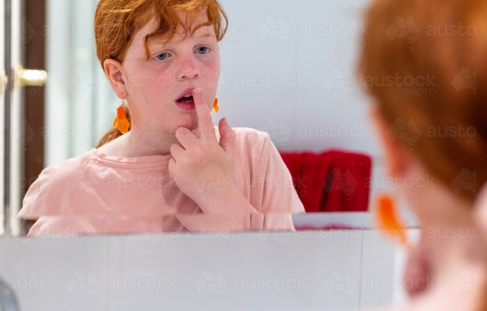 young girl looking in bathroom mirror putting lip balm on lips - Australian Stock Image