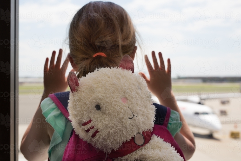 Young girl looking out the window at an airport with a toy cat - Australian Stock Image