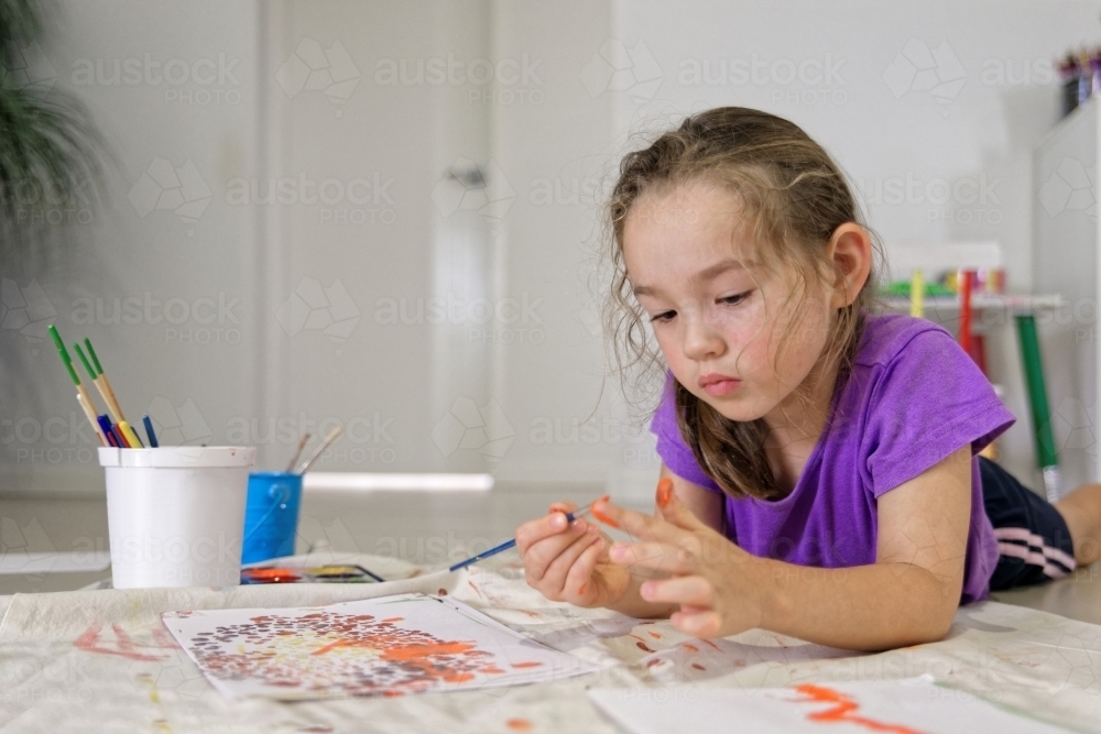 Young girl lying on the floor putting colour on her finger for painting a dot picture - Australian Stock Image
