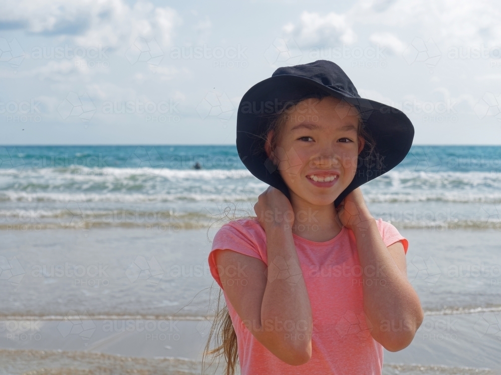 Young girl of mixed race holding her hat smiling on the beach - Australian Stock Image