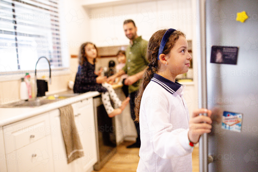 Young girl opening the fridge in the kitchen with dad and siblings in the background - Australian Stock Image