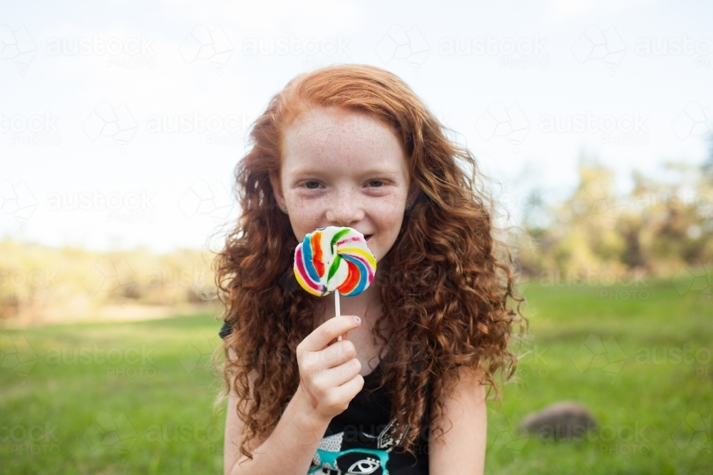 Young girl outside eating a rainbow lollipop - Australian Stock Image