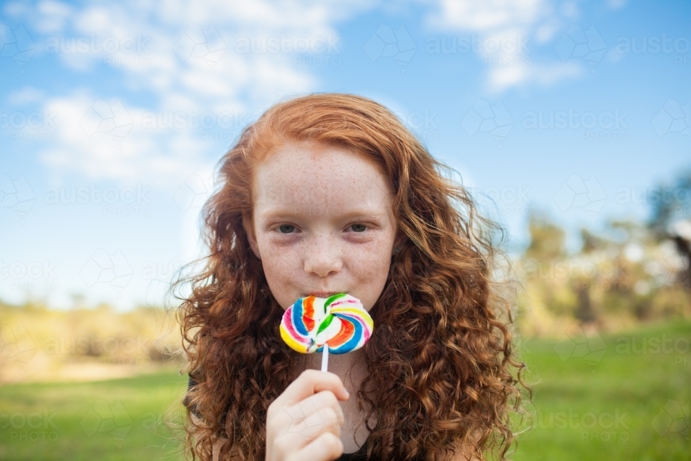Young girl outside eating a rainbow lollipop - Australian Stock Image