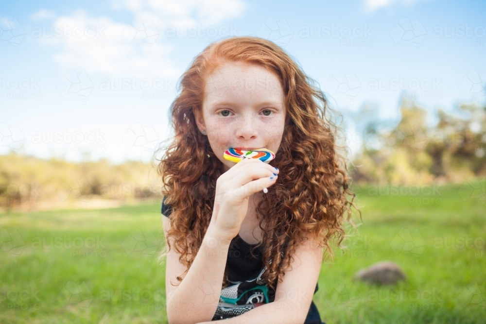 Young girl outside eating a rainbow lollipop - Australian Stock Image