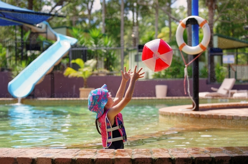 Young girl playing in the swimming pool wearing a swim vest throwing a ball - Australian Stock Image