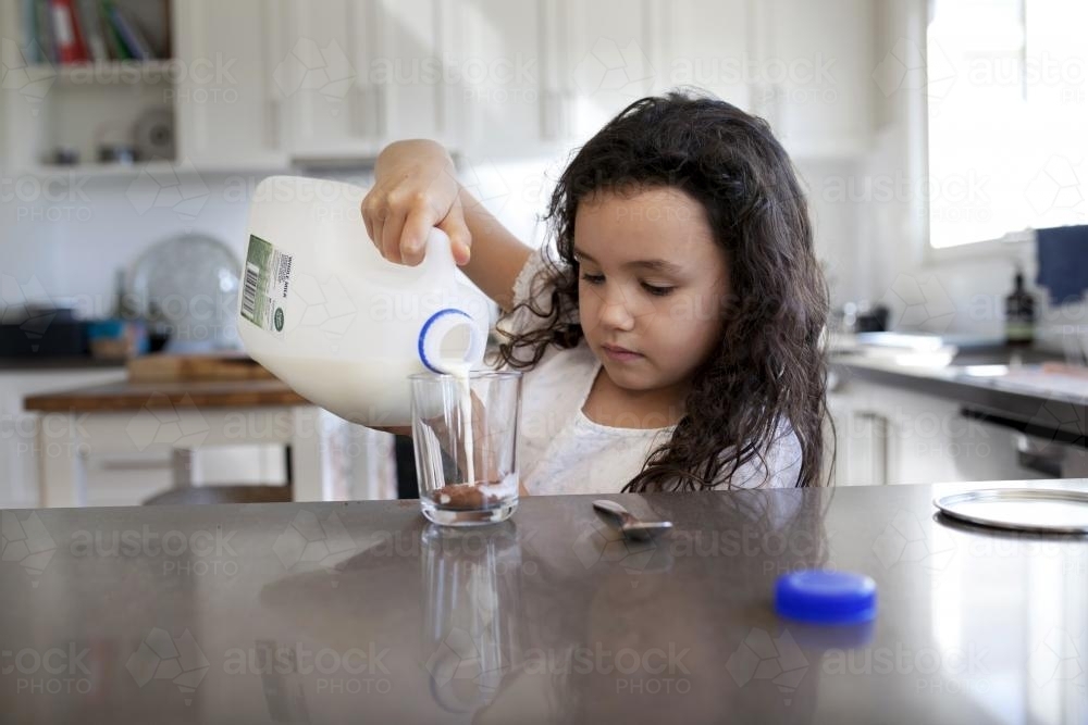 Young girl pouring milk into a glass in kitchen to make chocolate milk - Australian Stock Image