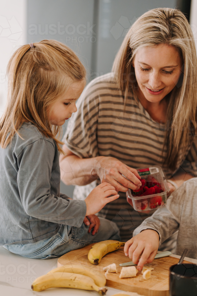 Young girl sits on the countertop while they prepare food in the kitchen. - Australian Stock Image