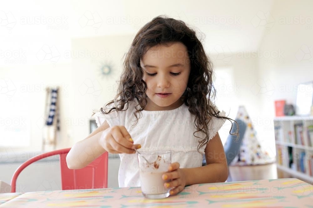 Young girl sitting at table mixing chocolate milk in a glass - Australian Stock Image