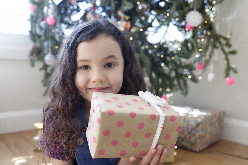 Young girl sitting in front of christmas tree holding a present up in front of her face - Australian Stock Image