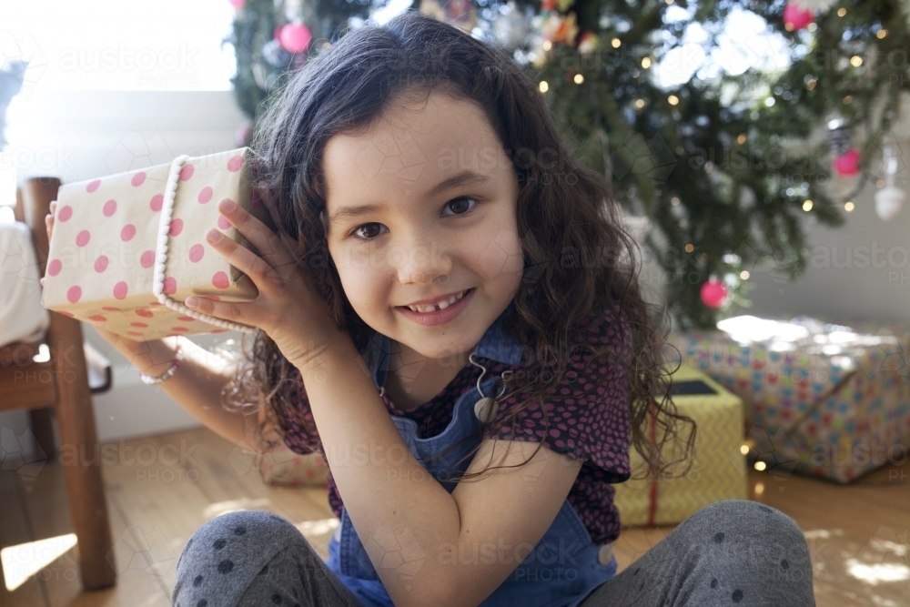 Young girl sitting in front of christmas tree holding a present up to her ear - Australian Stock Image