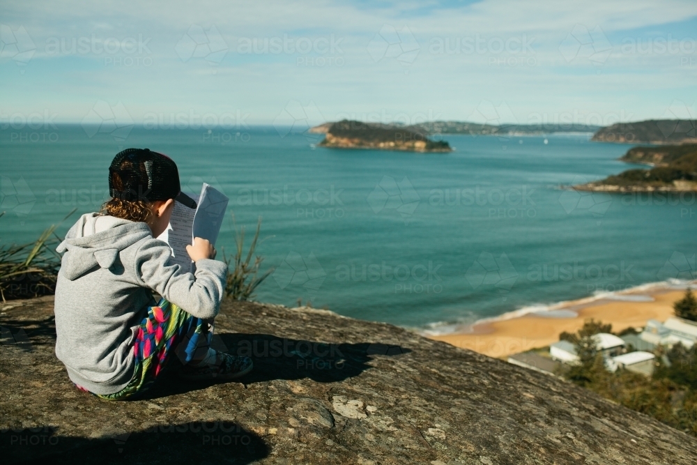 Young girl sitting on a rock overlooking the beach reading a book - Australian Stock Image