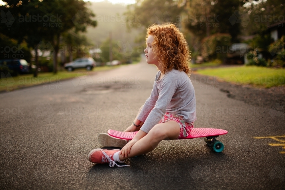 Young girl sitting on a skateboard on a road - Australian Stock Image