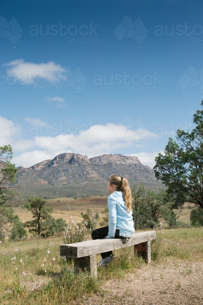 Young girl sitting on a wooden bench looking at mountains - Australian Stock Image