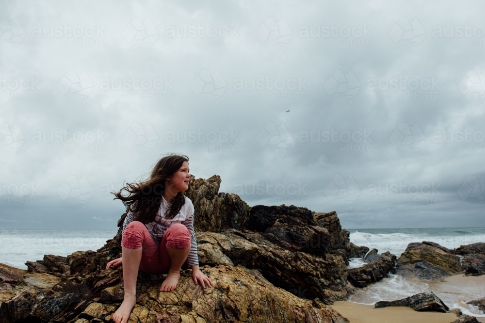 Young girl sitting on rocks at beach, wind blowing her hair - Australian Stock Image
