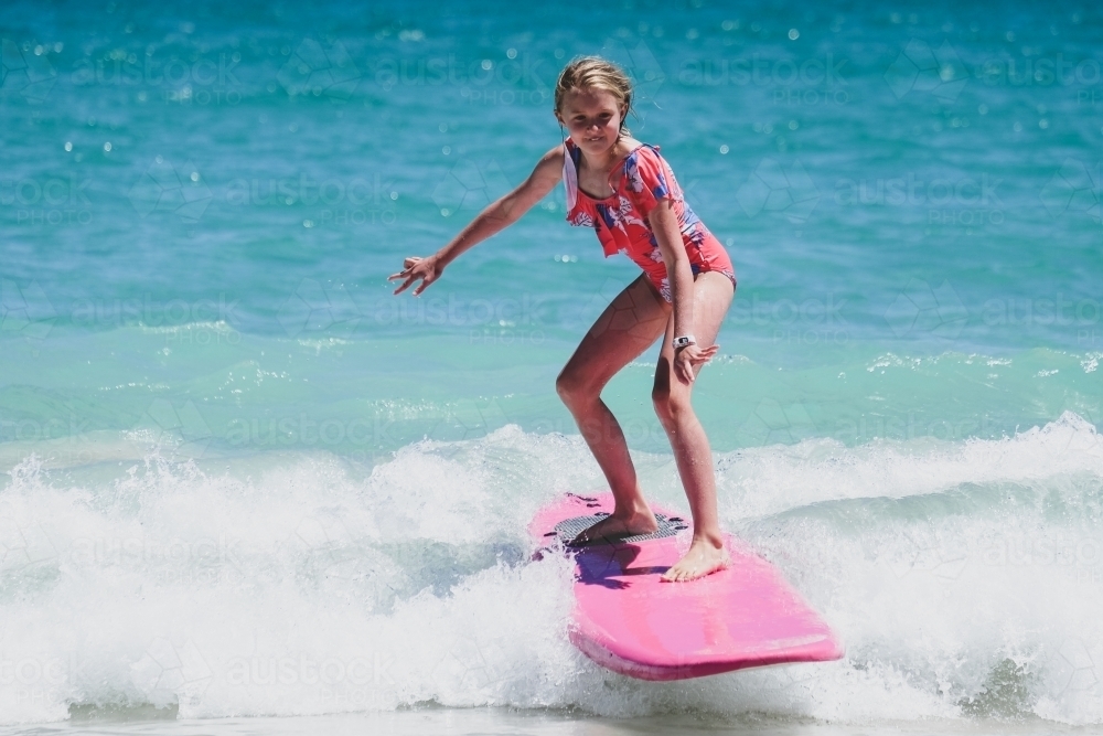 Young girl standing on pink soft board surfing the white wash waves wearing bathers - Australian Stock Image