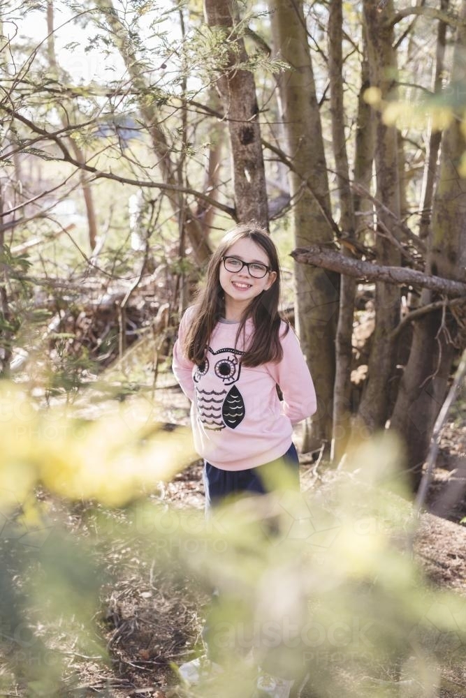 Young girl standing with arms behind back in some trees - Australian Stock Image