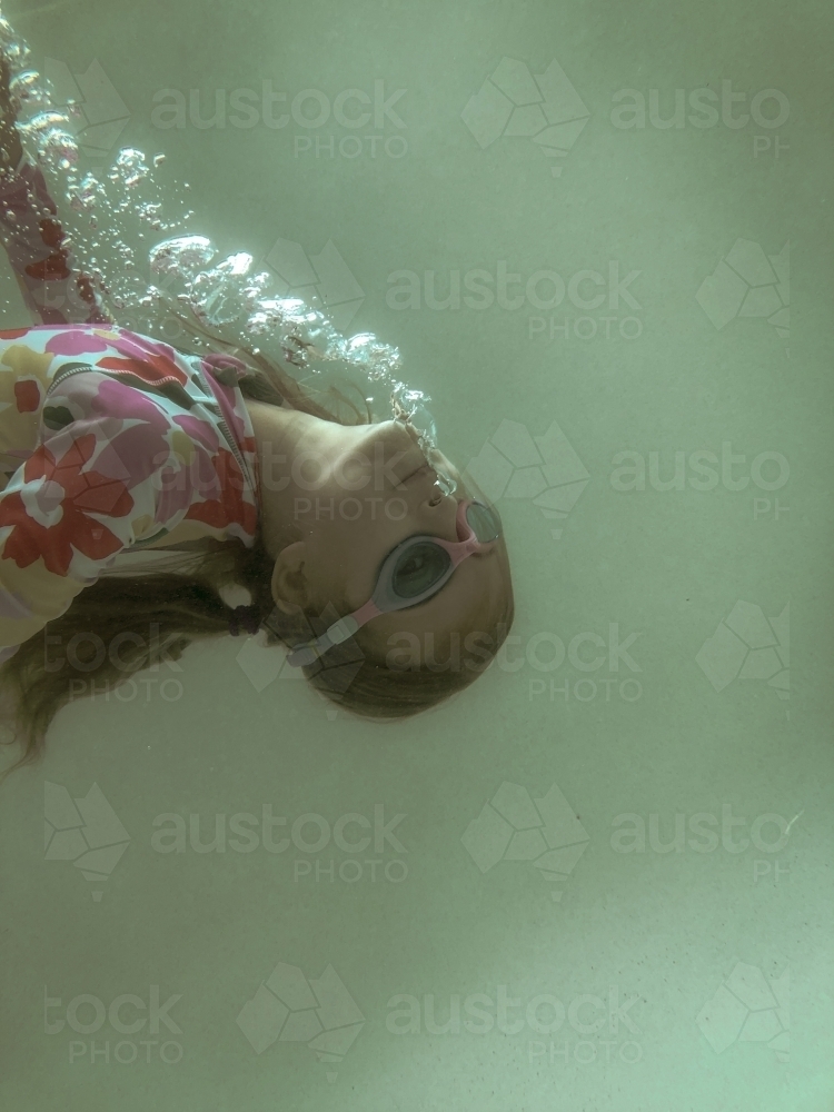 Young girl swimming underwater in pool wearing floral swim suit and goggles blowing bubbles - Australian Stock Image