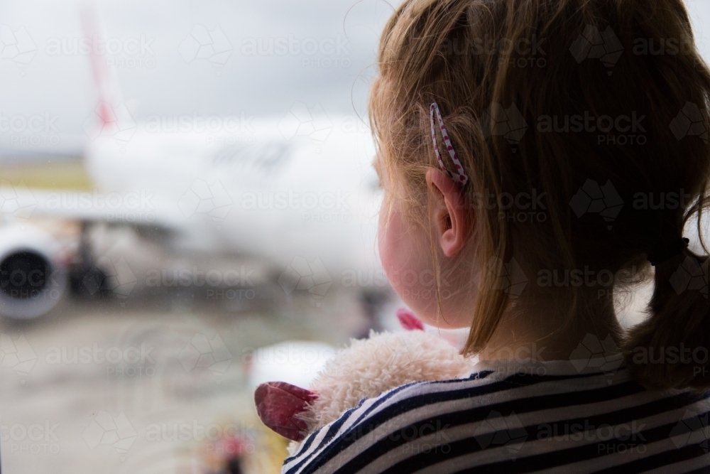 Young girl waiting for a flight, looking out a window while holding a toy cat - Australian Stock Image
