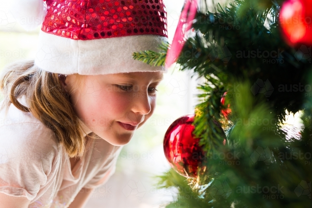 young girl wearing a santa hat smiling with a christmas tree - Australian Stock Image
