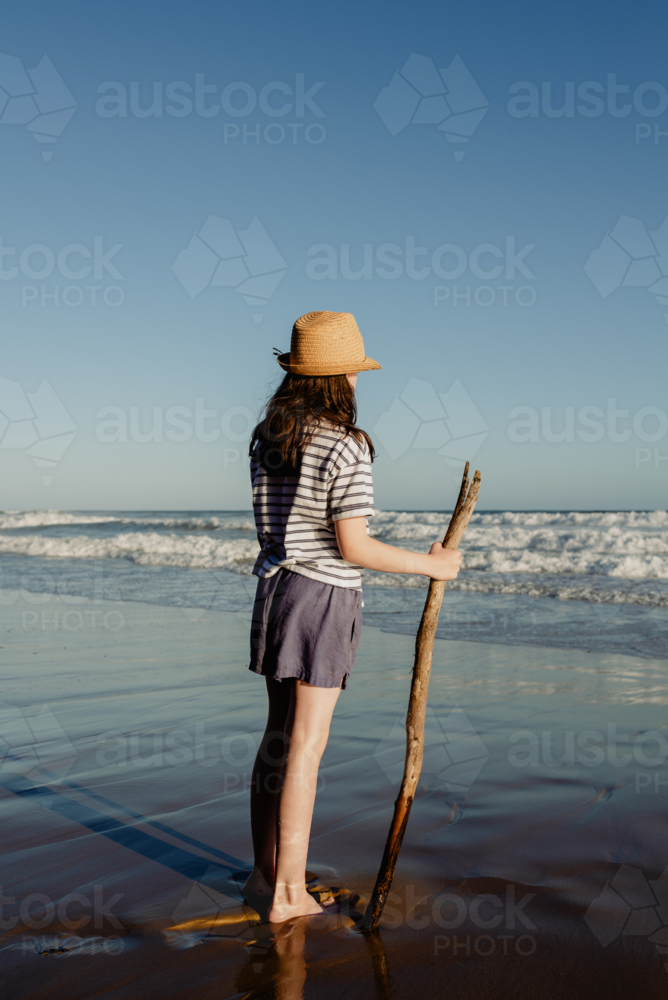 Young girl wearing a sunhat holding a stick on a beach in the afternoon - Australian Stock Image