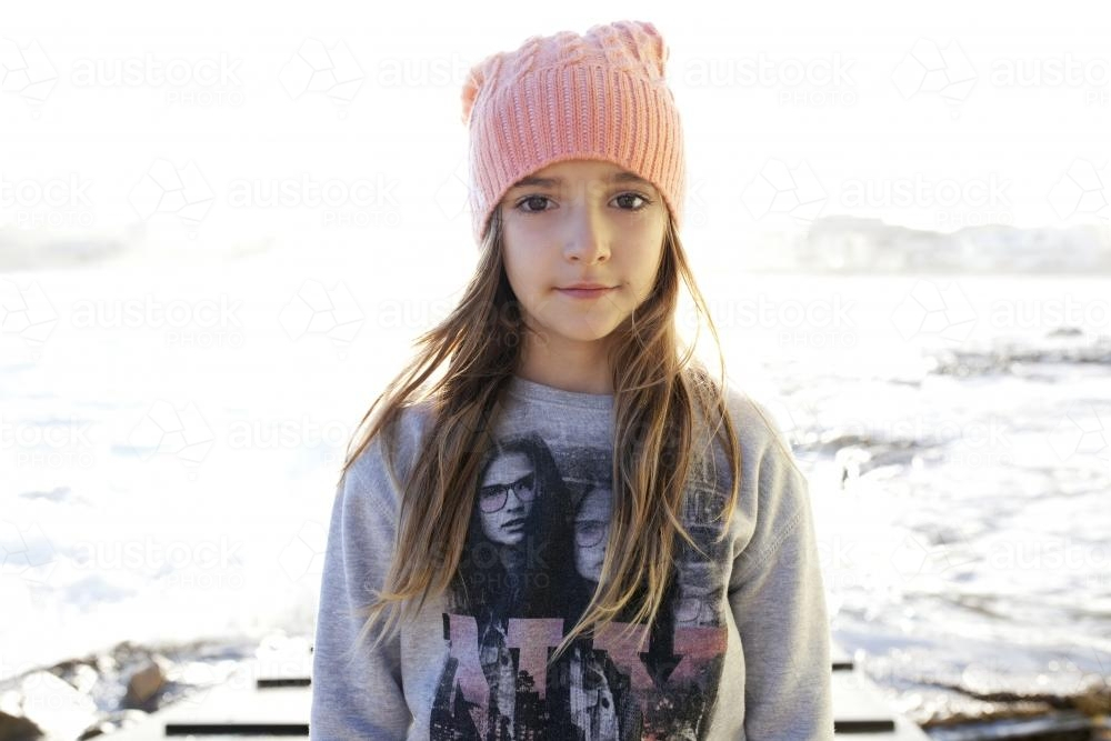 Young girl wearing beanie standing outdoors with beach behind her - Australian Stock Image