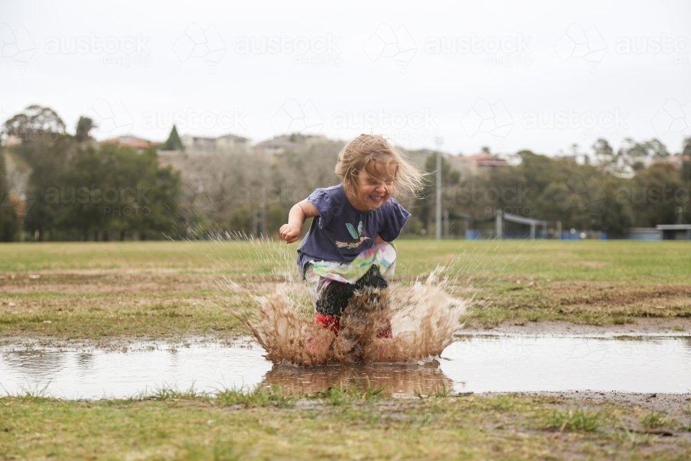 Young girl wearing red gumboots jumping in a rain puddle at a park - Australian Stock Image
