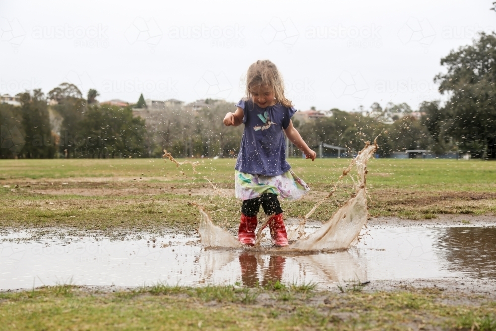 Young girl wearing red gumboots jumping in rain puddles - Australian Stock Image