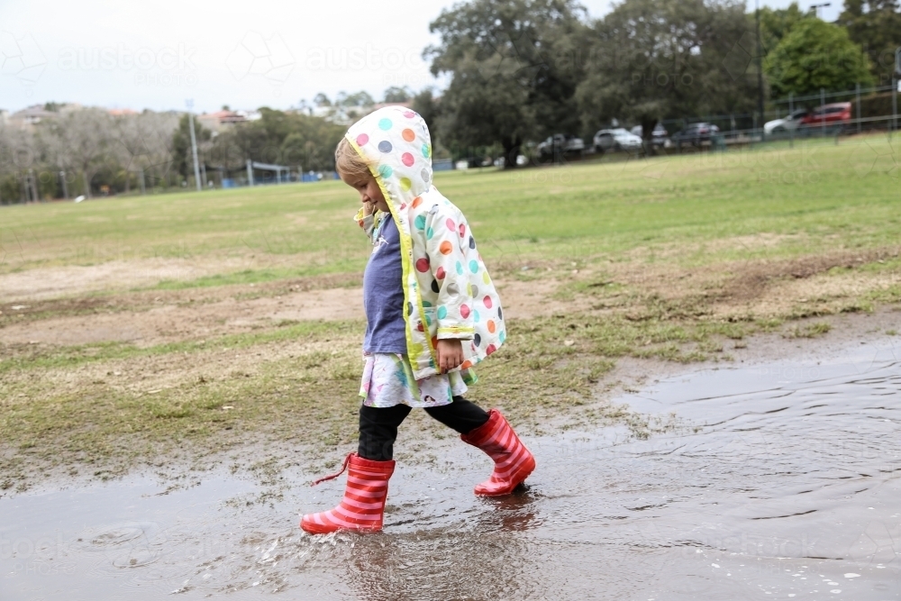 Young girl wearing spotted rain jacket playing in puddles - Australian Stock Image