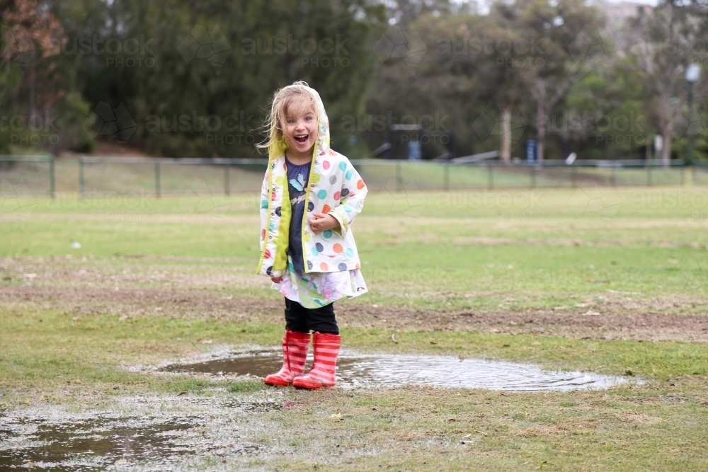 Young girl wearing spotted rain jacket playing in puddles - Australian Stock Image