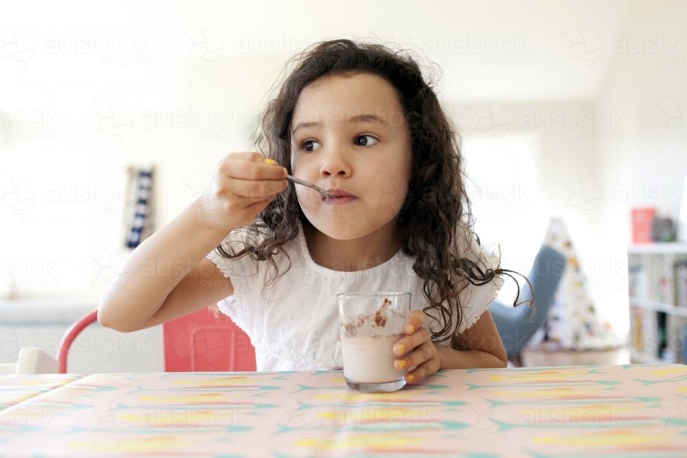 Young girl with a glass of chocolate milk sitting at table eating from spoon - Australian Stock Image