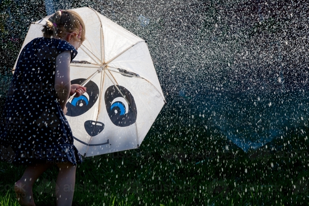 Young girl with an umbrella plating under the sprinkler - Australian Stock Image