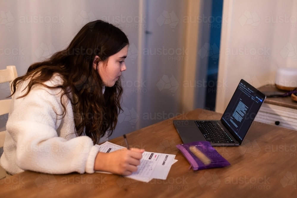 Young girl writing down notes while looking at her laptop computer on the table - Australian Stock Image