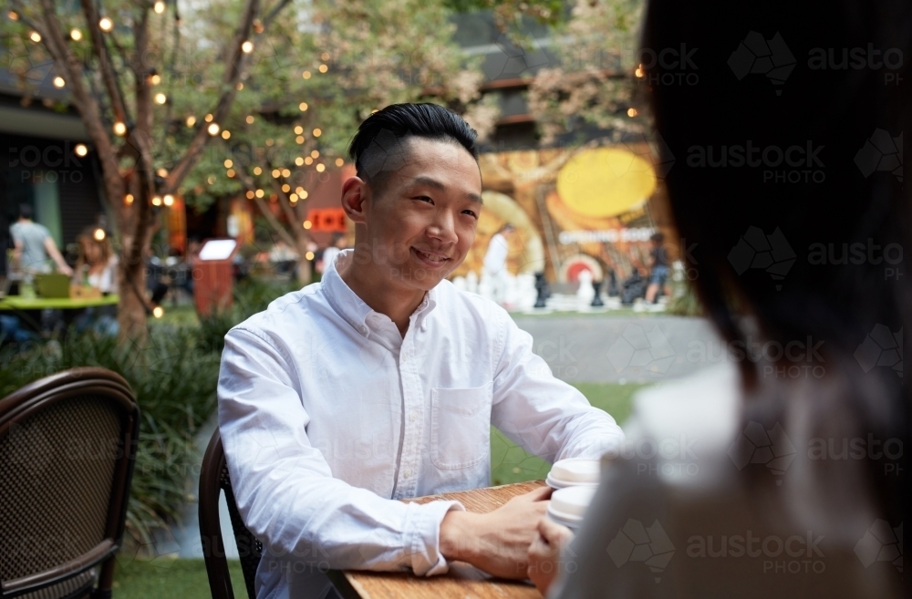 Young happy Asian man seated at outdoor restaurant with partner - Australian Stock Image