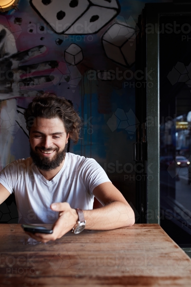 Young hipster man checking mobile phone and smiling at bar - Australian Stock Image
