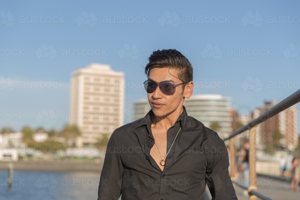 Young hispanic man wearing black shirt and sunglasses in trendy seaside setting - Australian Stock Image