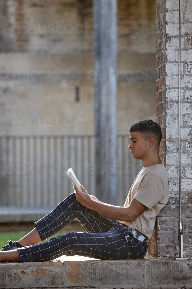 Young Indigenous man sitting against brick wall reading book - Australian Stock Image