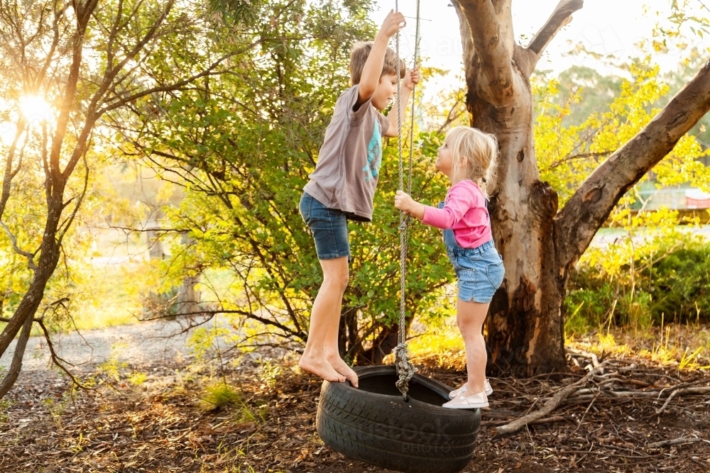 Young kids playing together on tire swing in backyard - Australian Stock Image