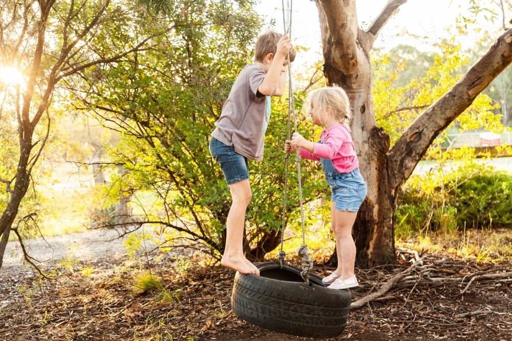 Young kids playing together on tire swing in backyard - Australian Stock Image