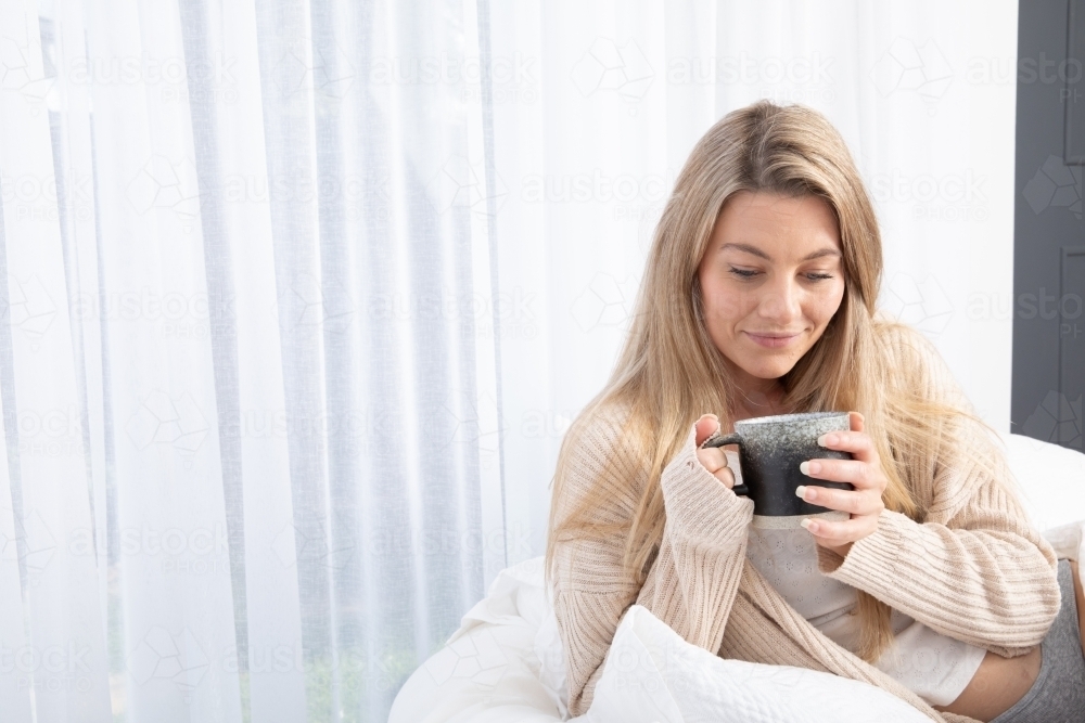 Young lady drinking hot drink in white bed relaxing - Australian Stock Image