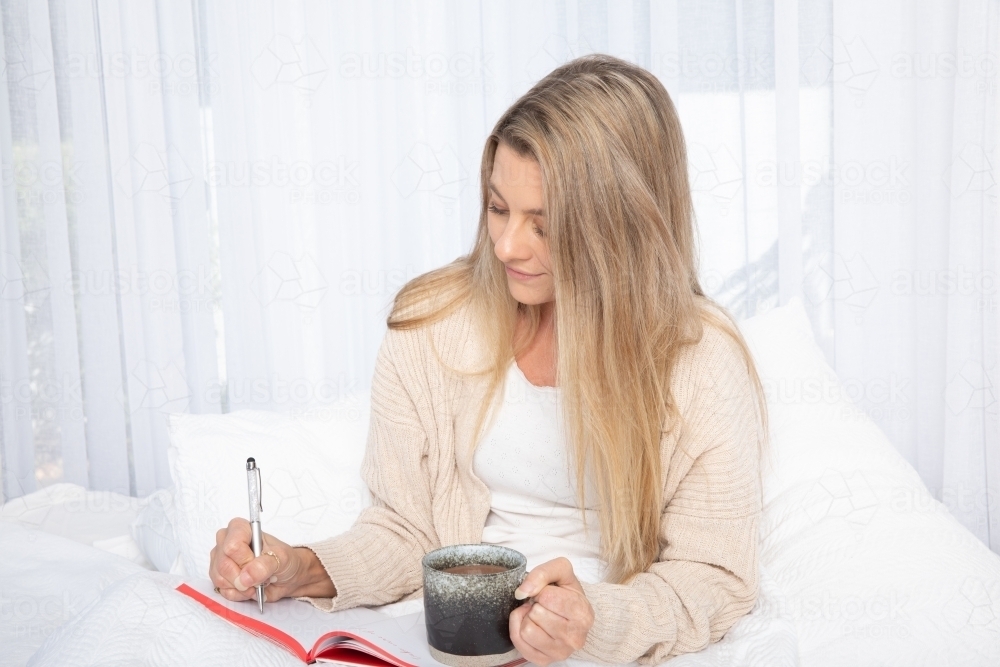 Young lady drinking hot drink in white bed writing in a journal - Australian Stock Image
