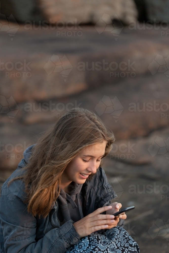 Young lady sitting on rocks and looking at smartphone at dusk - Australian Stock Image