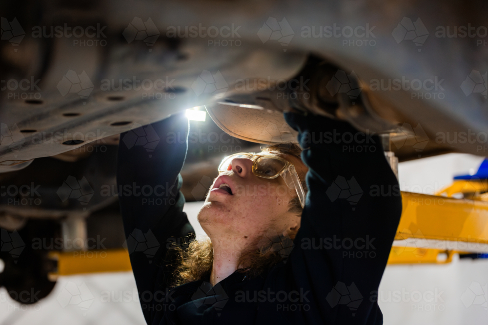 Young male mechanic working on the vehicles underbody with flashlight on - Australian Stock Image