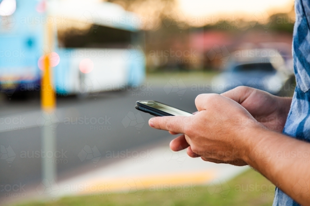 Young male person waiting at bus stop checking times on mobile phone app - Australian Stock Image