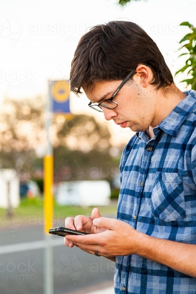 Young male person waiting at bus stop checking times on mobile phone app - Australian Stock Image