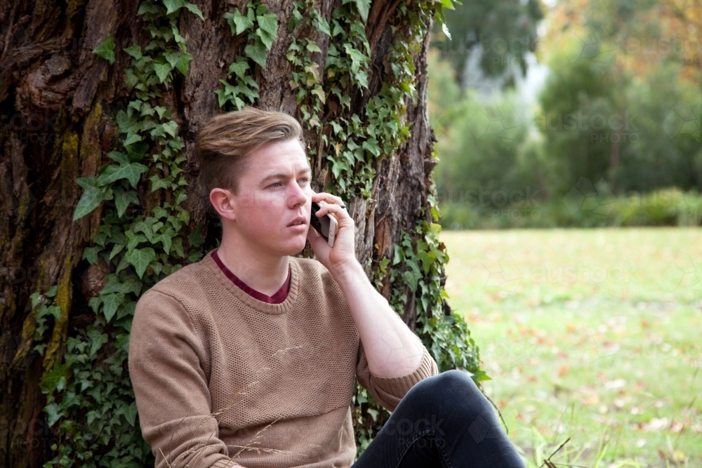Young male sitting outdoors talking on mobile phone - Australian Stock Image