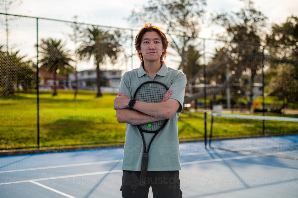 Young male stands confidently with a racquet on a tennis court in a park setting - Australian Stock Image