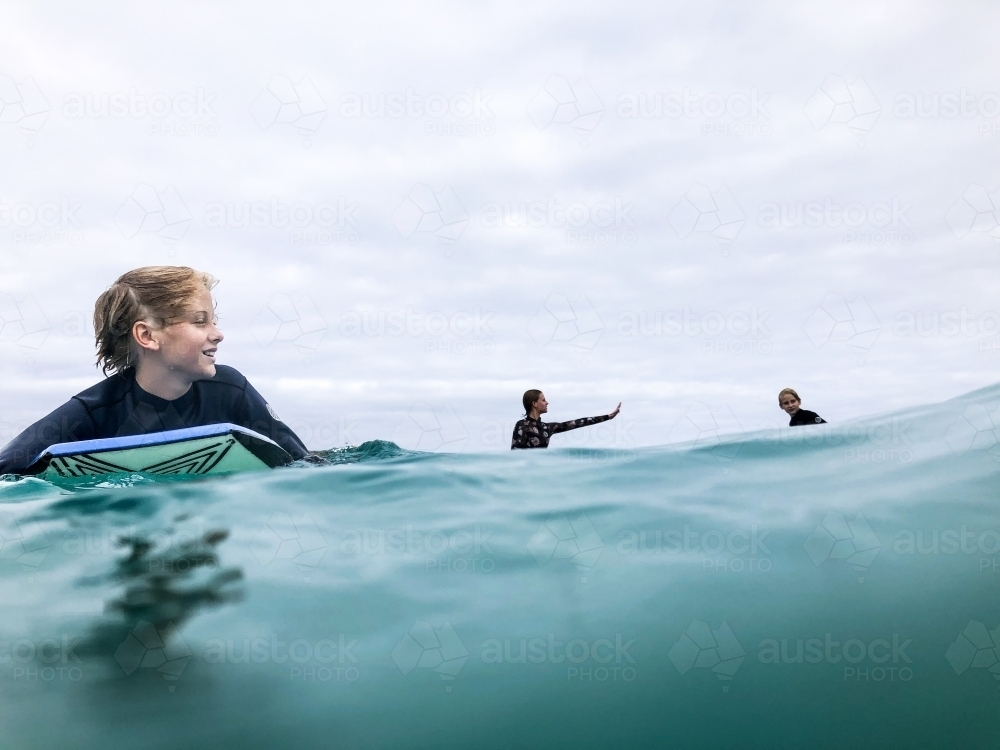 Young male surfer in ocean paddling on boogie board with two surfers sitting on boards in background - Australian Stock Image