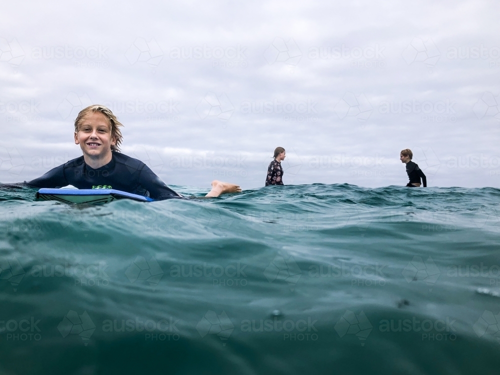Young male surfer in ocean paddling on boogie board with two surfers sitting on boards in background - Australian Stock Image
