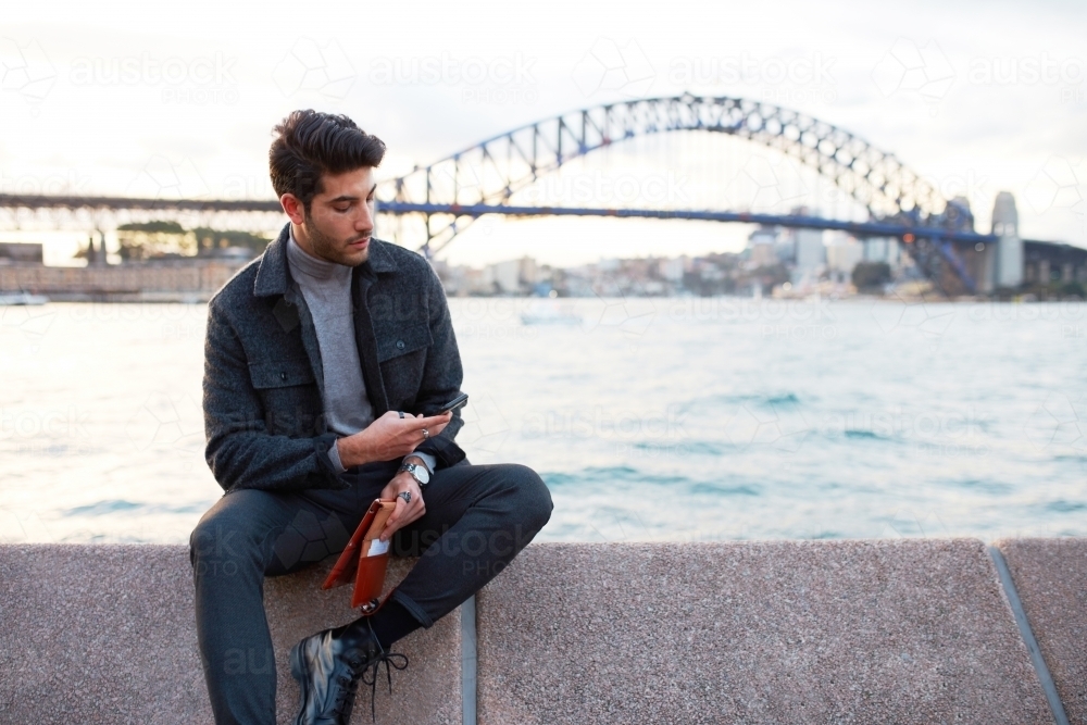 Young man checking phone in front of Sydney Harbour Bridge - Australian Stock Image