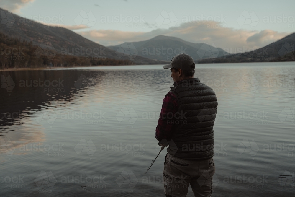 Young man fishing next to a calm lake with mountains in the background - Australian Stock Image